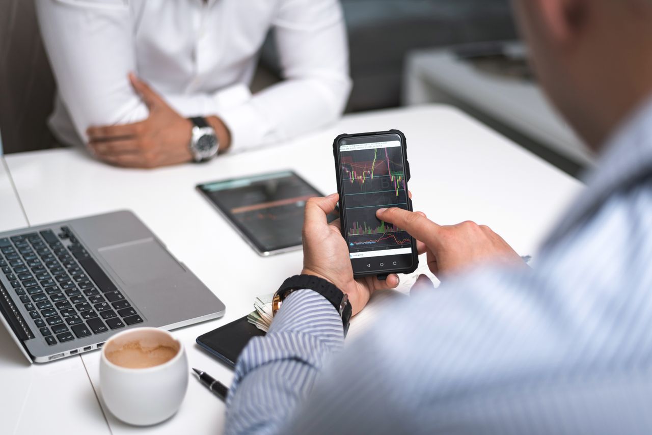 Two business professionals review stock market data on a smartphone and laptop in a contemporary office environment.